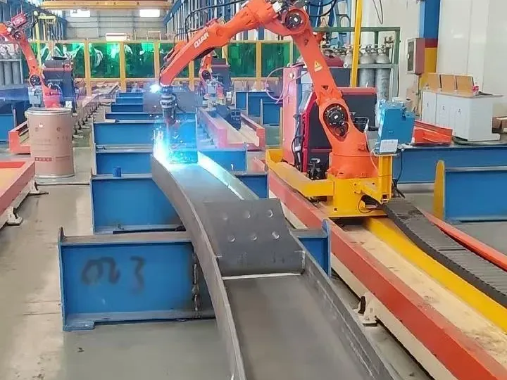 Robot naranja sobre riel soldando una viga curva de acero, con más estaciones detrás.
