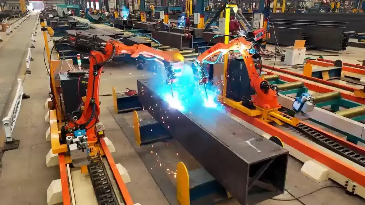 Dos robots naranjas sobre riel soldando una viga en cajón, con chispas en ambas antorchas.
