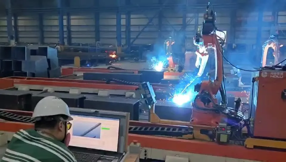 Operator in a hard hat monitoring welding software on a laptop beside working welding robots.