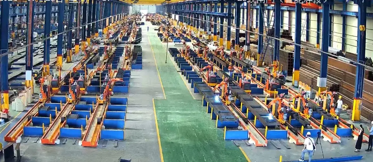 Long rows of welding robot stations along a green central aisle in a structural steel factory.
